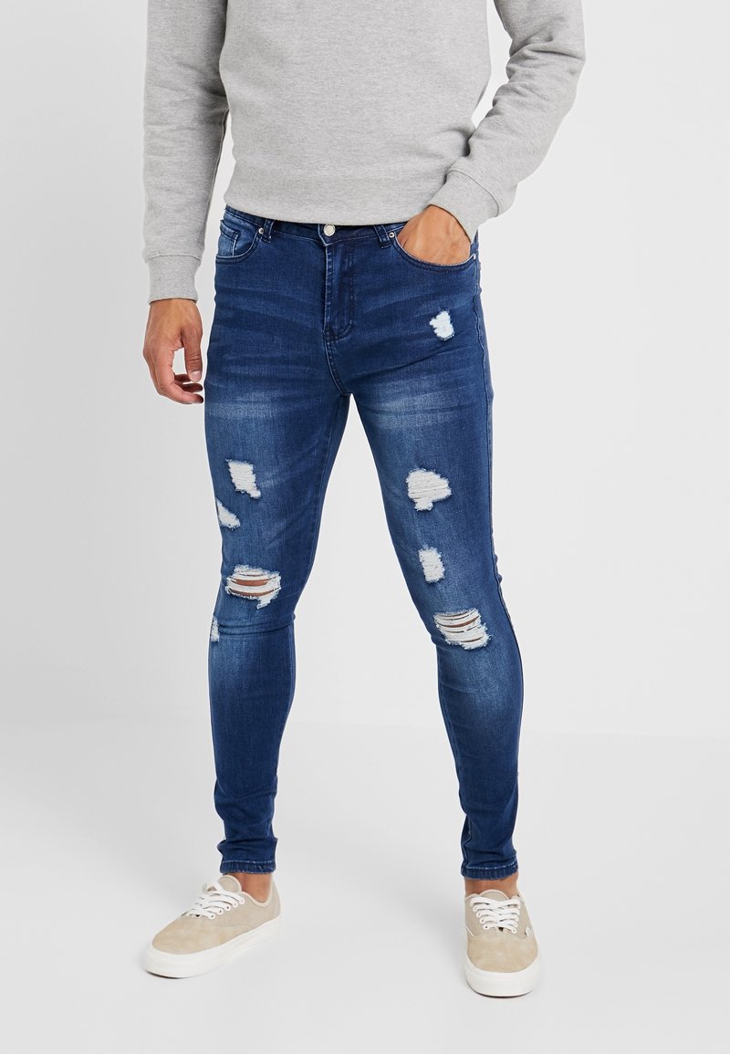 Jeans ajustados de color azul oscuro con desgastes y rotos, que cuentan con una textura de denim suave, cinco bolsillos y un cierre de botón. Combinados con zapatos beige.