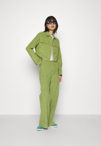 Chaqueta de pana verde con pantalones de pierna ancha a juego; presenta dos bolsillos en el pecho y un tejido texturizado; combinada con zapatillas de deporte en color azul verdoso y gafas de sol.