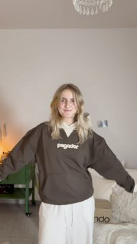 Brown oversized sweatshirt with a cream logo "pagador," features a hood and front pocket, paired with white trousers, in a light room.