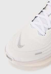 White Nike sneaker with a textured upper, prominent white laces, and grey accents, featuring a visible air unit in the sole.