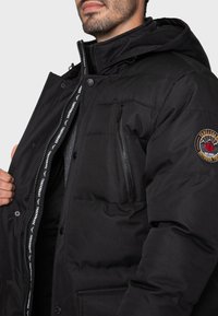 Giacca nera imbottita con tasca sul petto con zip, bottoni a pressione e cappuccio. Presenta una toppa con logo sulla manica e fodera interna a contrasto.