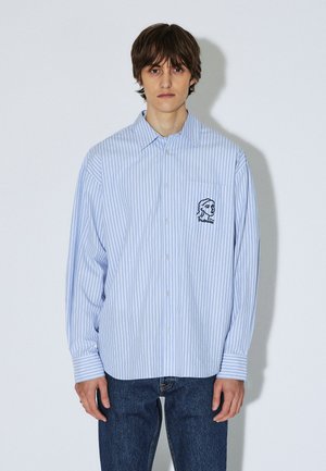 Vaaleansininen ja valkoraidallinen nappipaita, jossa on pitkät hihat ja rintatasku, jossa on kirjailtu profiilikuva. Puuvillakangas.