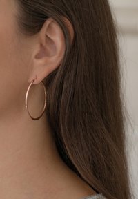 Pendiente de aro en oro rosa con un diseño circular delgado y un acabado pulido, colgando de una oreja perforada, complementando el cabello largo.
