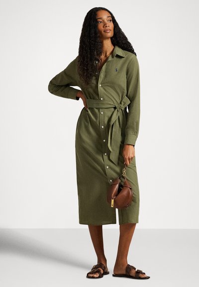 Olivno zelena obleka s srajčnimi rokavi, z gumbi spredaj in pasom v pasu. Gladek material, dolžina do kolen, kombinirana s rjavo torbico in sandali.