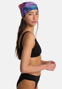 Jonge vrouw met lang donker haar, gekleed in een zwarte bikini en een kleurrijke paarse hoofddoek met patroon, kijkt over haar schouder tegen een witte achtergrond.