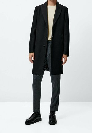 Czarny wełniany płaszcz z klapami z wycięciem, zapinany na jeden rząd guzików, zestawiony z szarymi spodniami o fakturze oraz czarnymi skórzanymi butami.