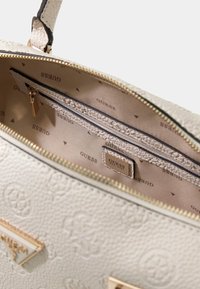 Borsa color crema con cerniera in tonalità oro. L'interno foderato presenta un delicato motivo "GUESS" e un'etichetta con logo in metallo.