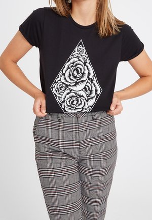 Kobieta ubrana w czarny t-shirt z białym wzorem kiści róż w kształcie diamentu, w zestawieniu z szarymi, kratowanymi, wysokowarstwowymi spodniami.
