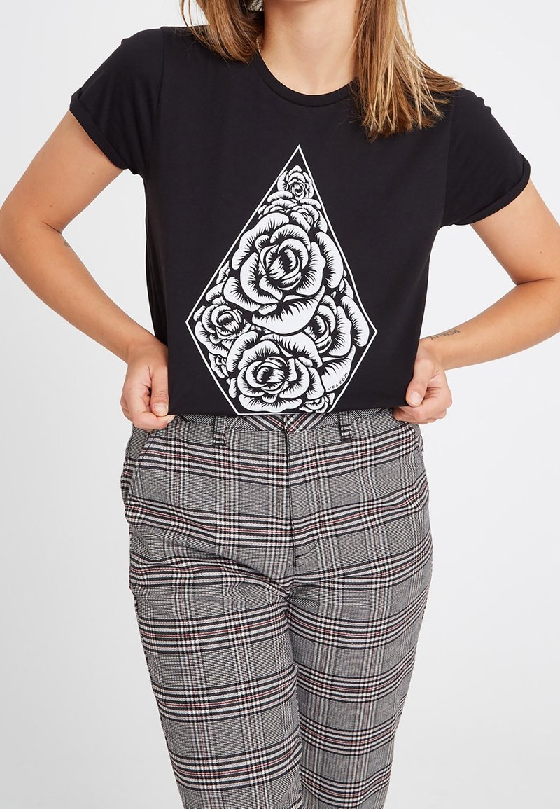 Femme portant un t-shirt noir avec un motif de bouquet de roses blanches en forme de losange, associé à un pantalon gris à carreaux taille haute.