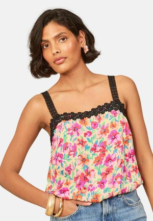 Vrouw draagt een mouwloze bloemen top met zwarte kanten afwerking, spijkerbroek, bloemvormige oorbellen en gouden armbanden om haar pols.