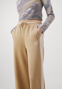 Pantalones de chándal beige con cintura elástica y cordón, que cuentan con bolsillos laterales y un logo blanco de Puma. El material parece suave y texturizado.