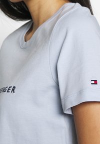 Vaaleansininen lyhythihainen t-paita, jossa on pyöreä kaula-aukko; siinä on tummansinisellä brodeerattu logo ja hihassa brändätty merkki punaisella, valkoisella ja sinisellä.