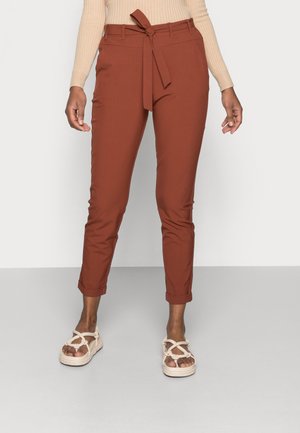 Femme portant un pantalon fuselé couleur rouille avec une ceinture à la taille et des ourlets retroussés, associé à des sandales beige à brides en corde.