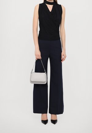 Mujer con top negro sin mangas y pantalones anchos azul marino, sosteniendo un bolso pequeño gris claro con cadena, y calzando tacones negros puntiagudos.