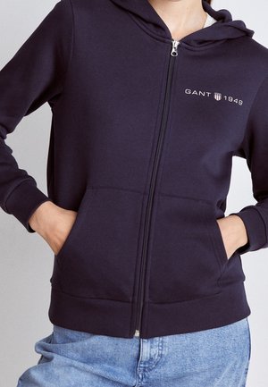Femme portant un sweat à capuche bleu marine zippé avec le logo "GANT 1949" sur la poitrine et un jean bleu clair, les mains dans les poches.