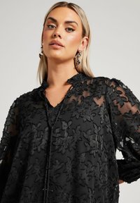 Blouse noire avec une broderie florale complexe, en tissu transparent, avec un décolleté en V et des manches longues. Accentuée par un détail à cordon.