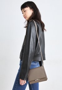 Zwarte leren jas met een getailleerd ontwerp, gecombineerd met een blauwe spijkerbroek. Een taupekleurige crossbodytas hangt aan één zijde, voorzien van een metalen sluiting.