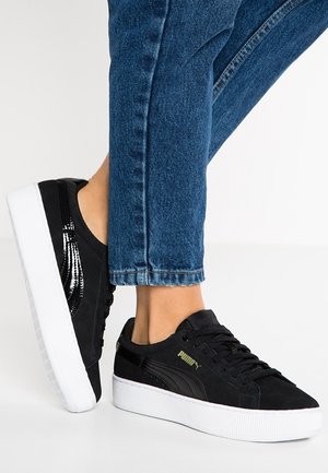 Personne portant des baskets Puma noires avec des semelles blanches épaisses et un jean bleu sur fond blanc.