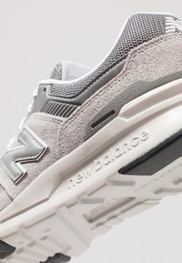 Nahaufnahme eines grauen und weißen New Balance Sneakers, der die Wildlederstruktur, ein Mesh-Obermaterial, ein gesticktes Logo und eine strukturierte Gummisohle mit Markennamen zeigt.