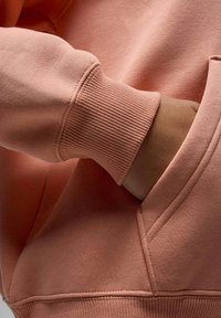 Sweatshirt rose clair avec des poignets côtelés et une poche. Le tissu semble doux, avec des détails de couture visibles et une coupe décontractée.