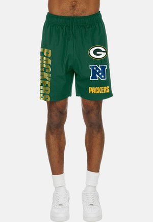 Grüne Sportshorts mit Green Bay Packers-Logos und Schriftzug, getragen von einer Person, die mit weißen Socken und weißen Turnschuhen steht.