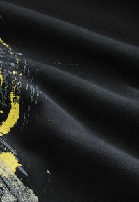 Melnas audums ar mīkstu tekstūru, kurā ir dzeltena un pelēka abstrakta dizaina kompozīcija, kas apvieno krāsu svītras un noplūdes, līdzīgu krāsas paklājumam.