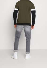 Camisa verde oliva con mangas largas negras y rayas blancas, combinada con jeans grises. Zapatos de caña alta blancos. Diseño casual y ajustado.