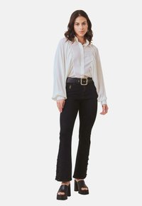 Camisa blanca de botones con detalles fruncidos y mangas largas, combinada con jeans negros de cintura alta con acentos laterales y sandalias negras gruesas.