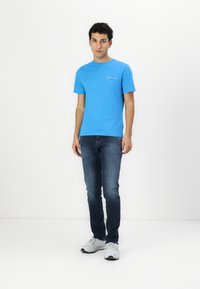 Ryški mėlynas medvilninis T-shirt su trumpomis rankovėmis ir apvaliu iškirpimu, derinamas su tamsiais džinsais ir pilkais sportiniais bateliais. Ant marškinėlių paprastas logotipo detalė.