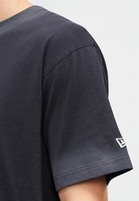 T-shirt de manga curta cinza escuro, feito de algodão suave. Apresenta um decote redondo e um detalhe em logotipo branco na manga. Textura suave.