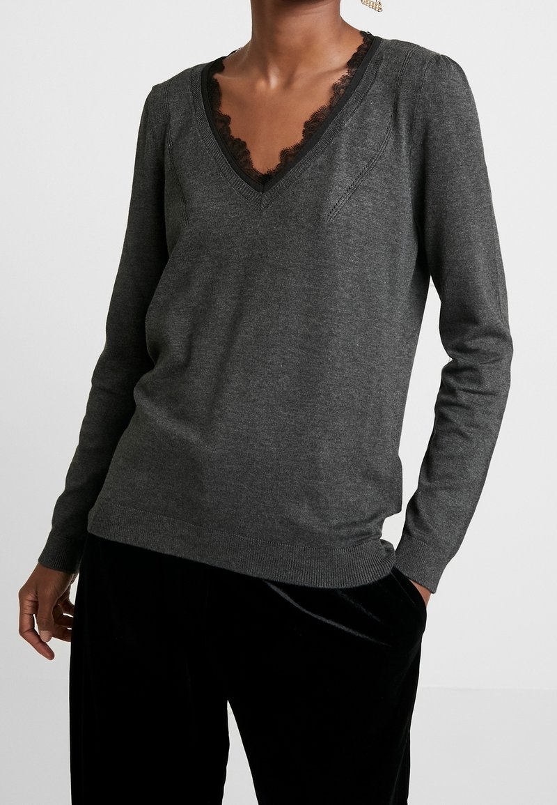 Pull à manches longues gris foncé avec col en V et bordure en dentelle noire porté par une personne avec une main dans la poche d'un pantalon en velours noir.