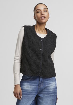 Nő fekete gombos polár mellényt viselnyi hosszú ujjú bézs felső és kék farmernadrág fölött, egyszínű világosszürke háttér előtt áll.