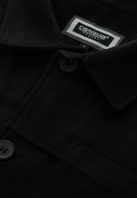 Giubbotto in suede nero con colletto a punta, due bottoni e un'etichetta con la scritta "CARISMA CASUAL". Texture liscia e design minimalista.