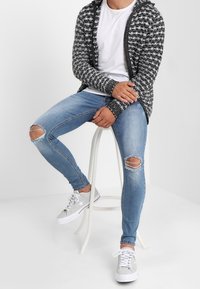 Cardigan a maglia bianco e nero sopra una t-shirt bianca, abbinato a jeans skinny strappati azzurri chiari e sneakers grigie, seduto su uno sgabello.