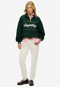Groene fleece pullover met witte logo, half-zip hals, en gestreepte mouwen, gedragen over een roze en witte gestreepte shirt en crèmekleurige broek.
