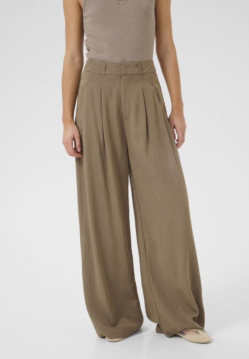 Femme portant un pantalon kaki taille haute, large et ample, et un top beige sans manches, debout les mains détendues le long du corps.
