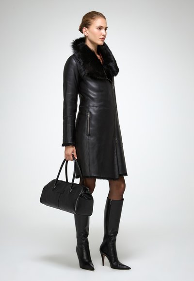 Donna che indossa un cappotto di pelle nera con colletto e polsini in pelliccia, tiene in mano una borsa di pelle nera e indossa stivali neri con tacco alti fino al ginocchio.