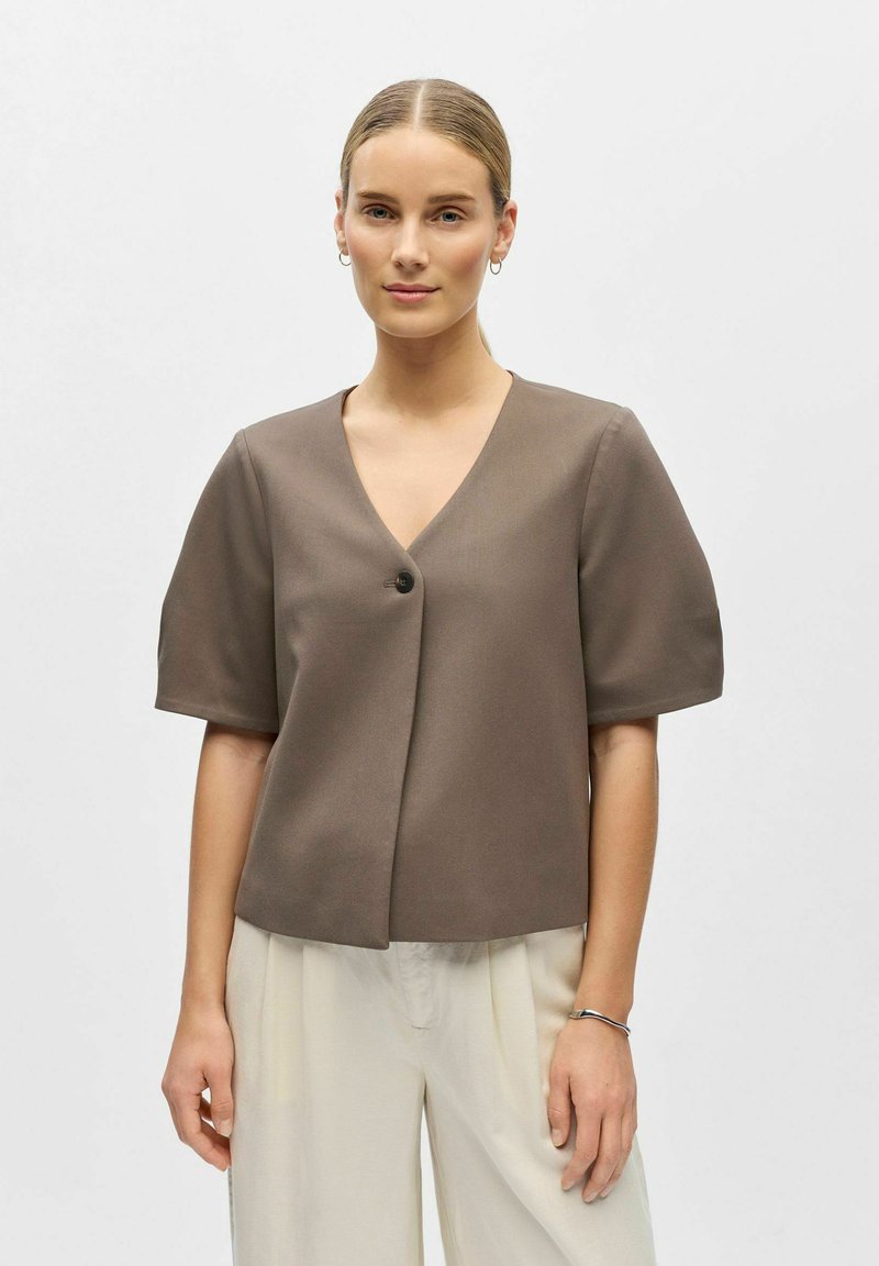 Blouse marron à manches courtes avec un col en V et une fermeture à un seul bouton. Texture lisse, coupe décontractée et design minimaliste.