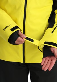 Die gelbe wasserdichte Jacke verfügt über einen Reißverschluss vorne, schwarze Akzente und verstellbare Ärmelbündchen mit Klettverschluss. Hände zeigen den Mechanismus zur Einstellung der Ärmelbündchen.