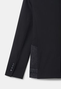 Manche de veste de costume noire avec quatre boutons noirs et une fente latérale laissant apparaître un tissu en denim gris foncé en dessous.