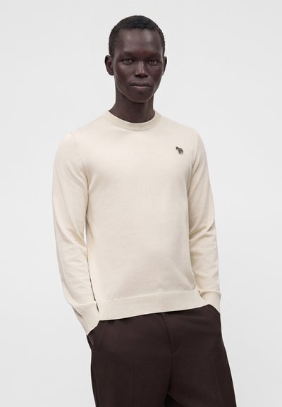 Homme portant un pull crème à manches longues avec un petit logo zèbre sur la poitrine et un pantalon marron foncé, debout les mains dans les poches.