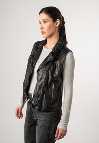 Gilet da motociclista in pelle nera con chiusura a zip asimmetrica, tasche con zip e una texture liscia. Indossato sopra una maglietta a maniche lunghe grigia chiara.