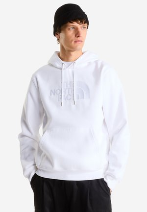 Giovane uomo che indossa una felpa bianca con cappuccio, tasca frontale e logo discreto "The North Face", berretto nero e pantaloni neri, che guarda di lato.