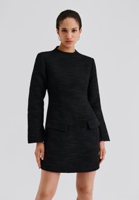 Schwarzes strukturiertes Kleid mit langen Ärmeln, taillierter Passform und zwei vorderen Taschen. Glattes Gewebe mit dezenten Mustern, knielanger Saum.