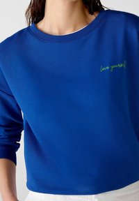 Persona que lleva una sudadera azul real con texto bordado en verde "ámate a ti mismo" en el pecho, combinada con pantalones blancos.