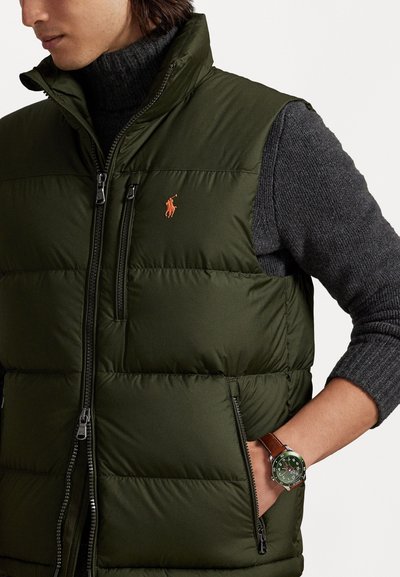 Chaleco acolchado verde oliva con cuello alto, frente con cremallera y acolchado horizontal. Presenta un pequeño logo bordado y detalles de bolsillos laterales.