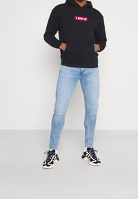 Sudadera con capucha negra con el logo rojo de Levi's, combinada con vaqueros ajustados azul claro y zapatillas negras y blancas. Fondo liso.