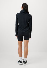 Camisola preta de mangas longas com capuz, de design ajustado e tecido texturizado. Combinada com calções pretos e ténis desportivos com pormenores em verde.