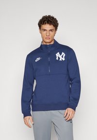 Námorníckomodrý Nike sveter so štvrtinovým zipsom, s bielym logom New York Yankees, ribovaný manžety a hladká textúra.