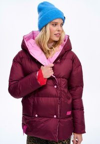 Bordowa kurtka puchowa z różowym wnętrzem, z zatrzaskami i wysokim kołnierzem. Połączona z niebieską dzianinową czapką i spodniami w zwierzęcy wzór.